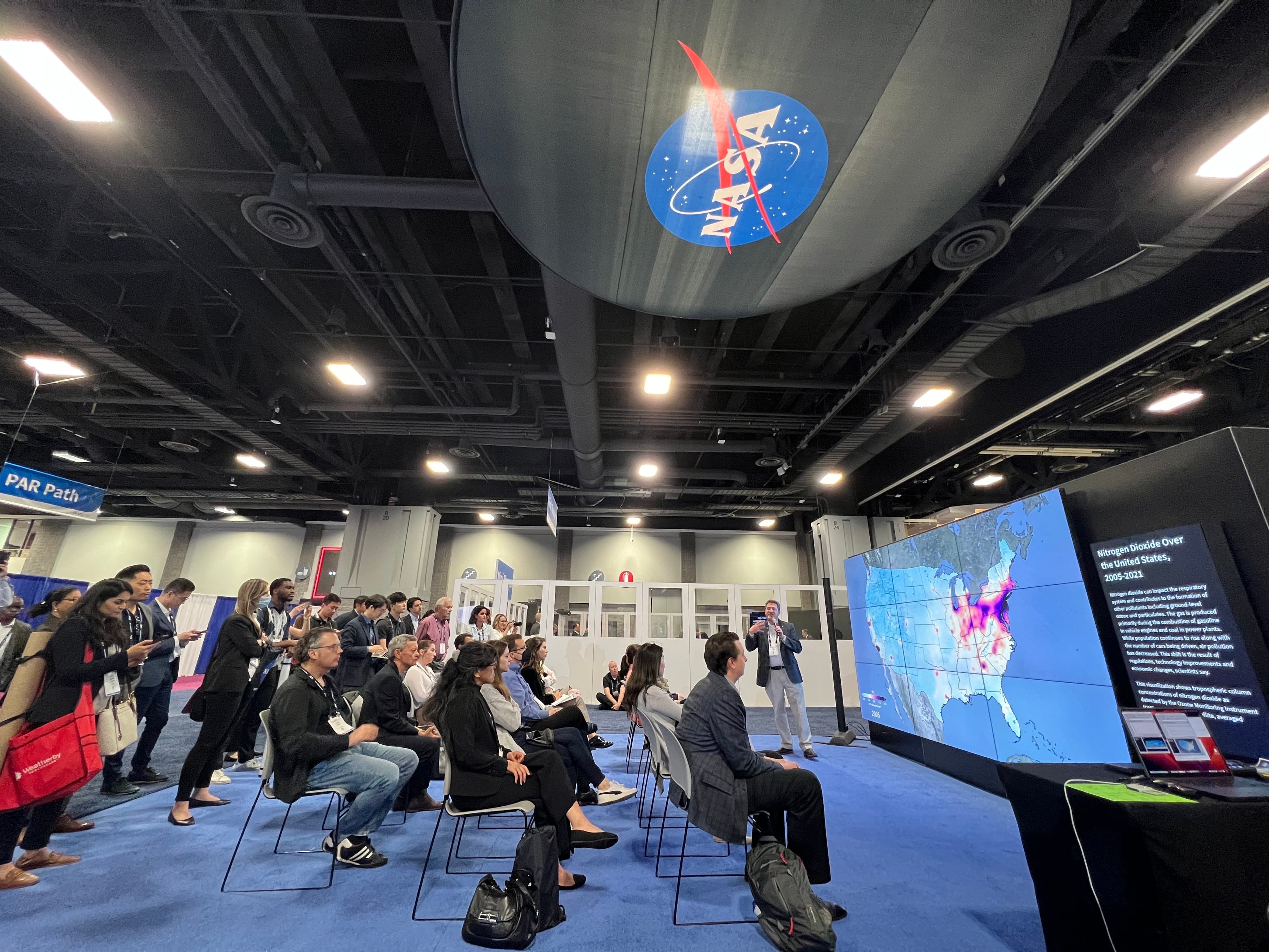 A seated crowd in front of a screen the size of 9 large televisions together. On the screen is a heatmap of the United States. On the ceiling is the NASA logo.